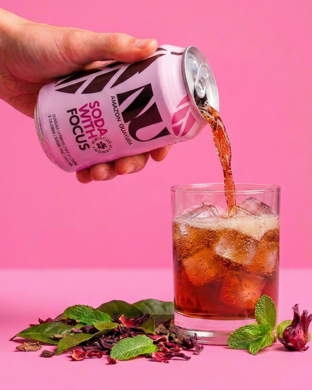 A hand holds a can of Soda with Focus, pouring the dark reddish-brown drink into a glass of ice. The background is a solid pink studio backdrop with dried flowers and mint leaves on the surface.