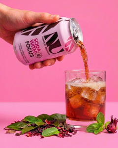 A hand holds a can of Soda with Focus, pouring the dark reddish-brown drink into a glass of ice. The background is a solid pink studio backdrop with dried flowers and mint leaves on the surface.